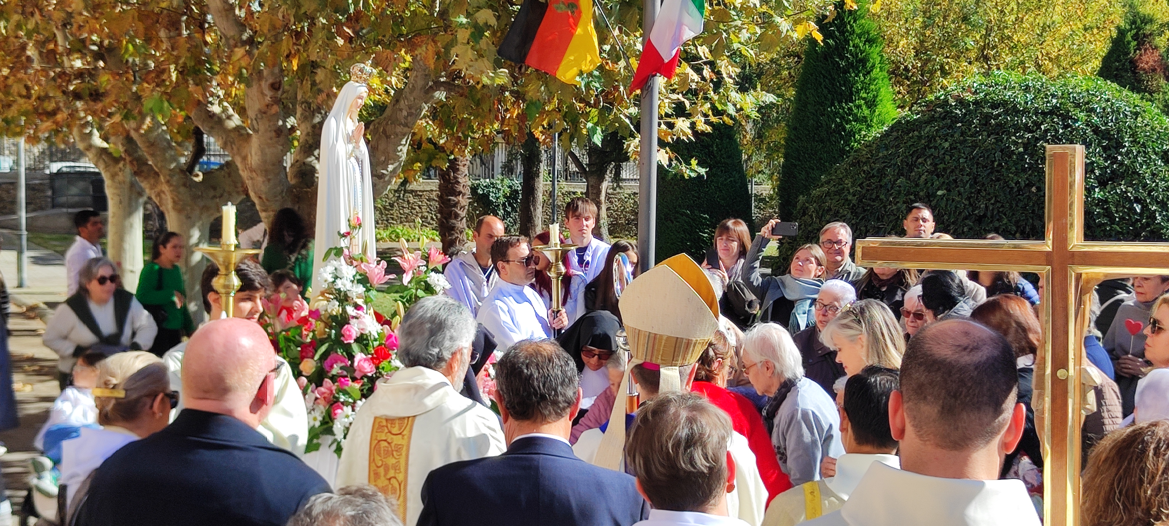 Cerimónia religiosa ao ar livre com a Imagem Peregrina de Nossa Senhora de Fátima rodeada de flores, membros do clero e muitos fiéis reunidos em oração.