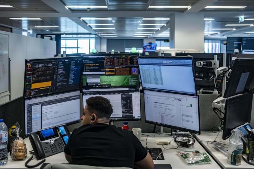 Pessoa sentada numa secretária com vários monitores a mostrar gráficos e dados financeiros, num escritório moderno, ilustrando o ambiente de análise económica e monitorização de mercados discutido no artigo sobre estagflação.