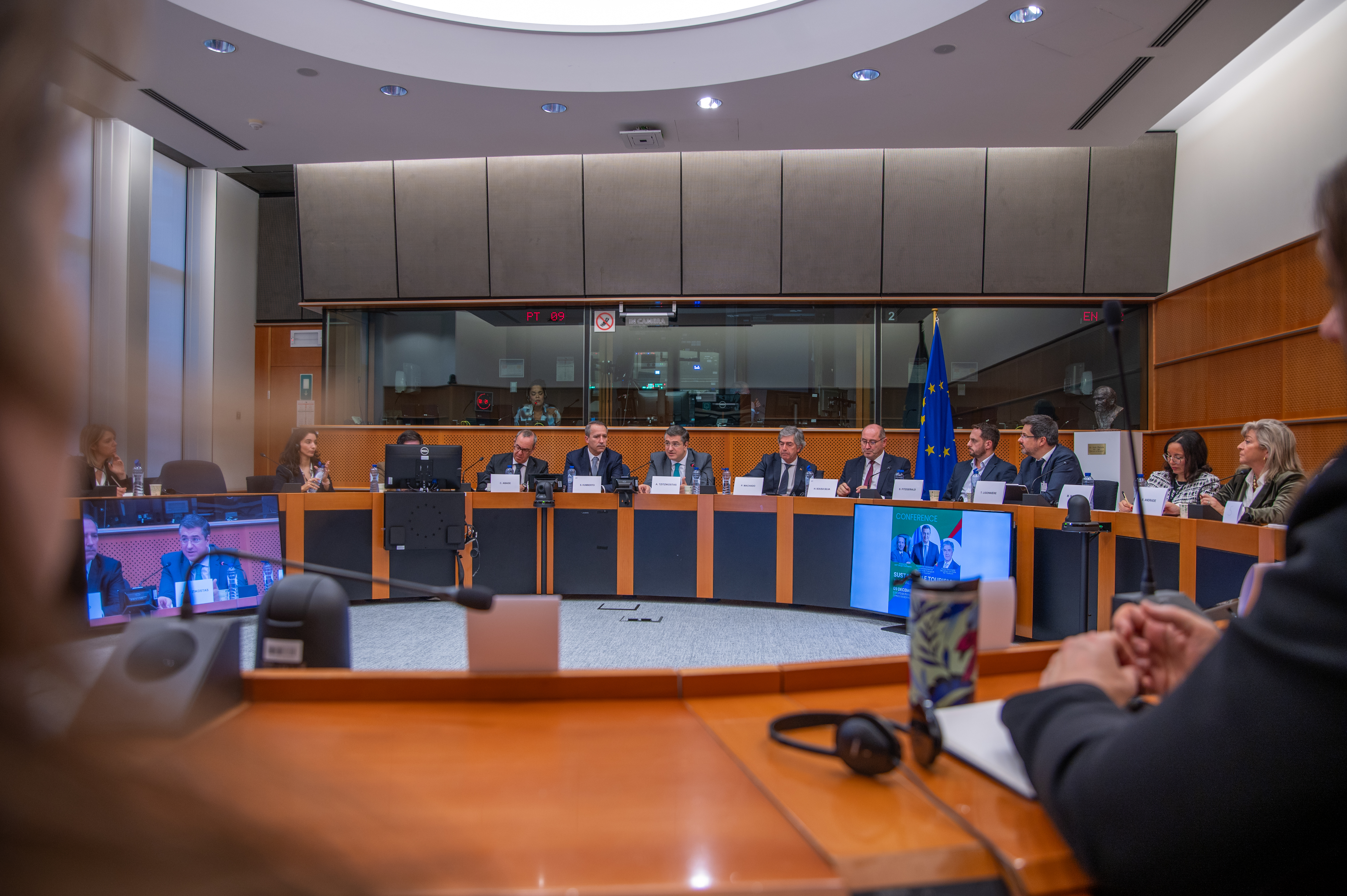 Sessão formal no Parlamento Europeu, com participantes sentados em torno de uma mesa em U, microfones e bandeira da União Europeia visíveis, ilustrando um debate sobre turismo sustentável.