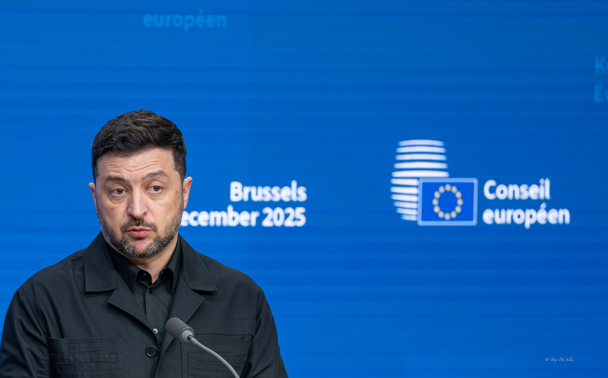 O Presidente da Ucrânia, Volodymyr Zelensky, fala num púlpito em Bruxelas, com o logótipo do Conselho Europeu e a data de dezembro de 2025 projetados num ecrã azul atrás dele.