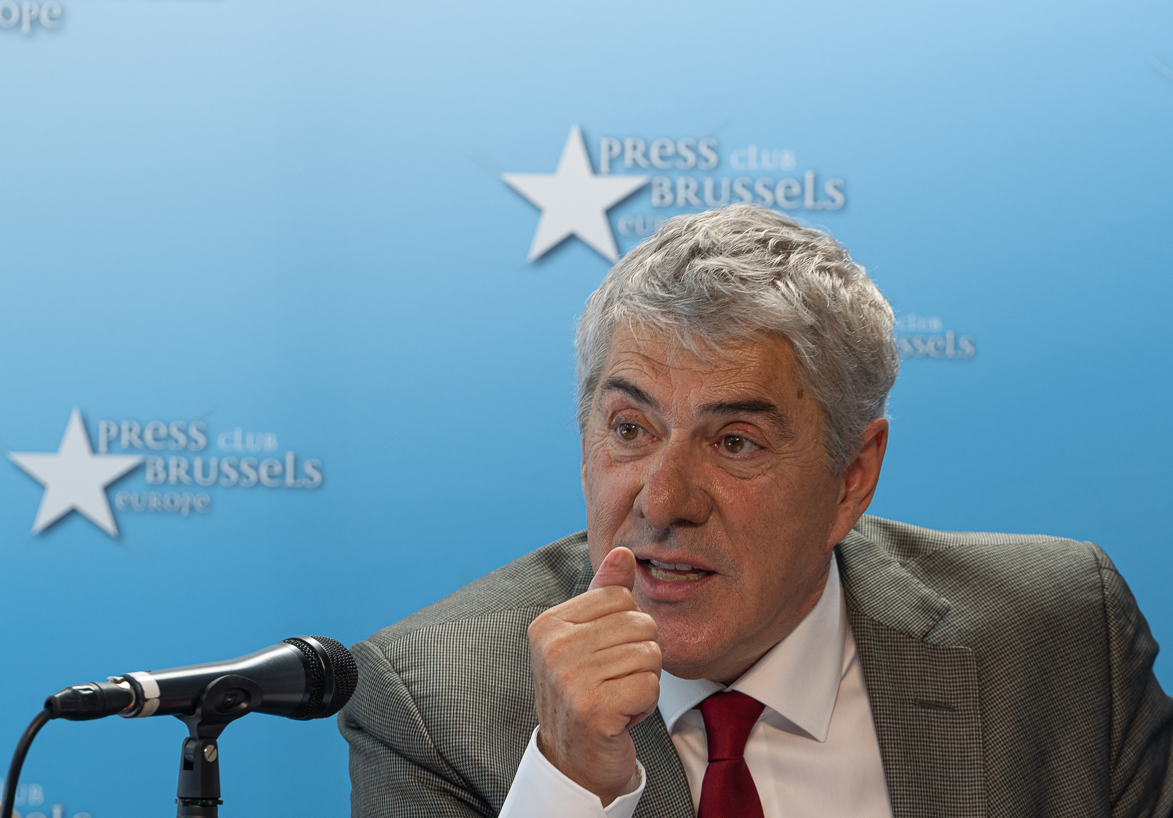 Homem de fato e gravata a falar num microfone, sentado em frente a um fundo azul com o logótipo 'Press Club Brussels Europe', sugerindo uma conferência de imprensa ou declaração pública.