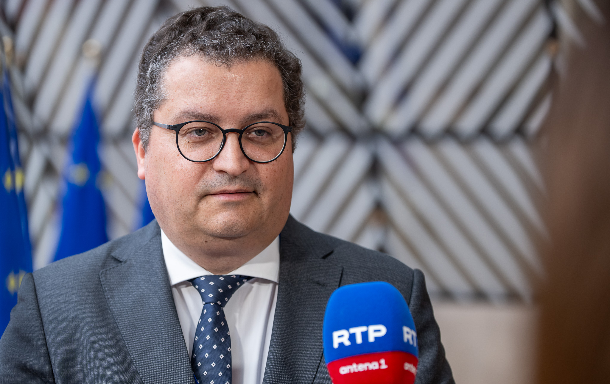 O ministro das Finanças português, de fato e gravata, fala à imprensa em Bruxelas, com microfones de canais portugueses à sua frente e bandeiras da União Europeia e outros países ao fundo.