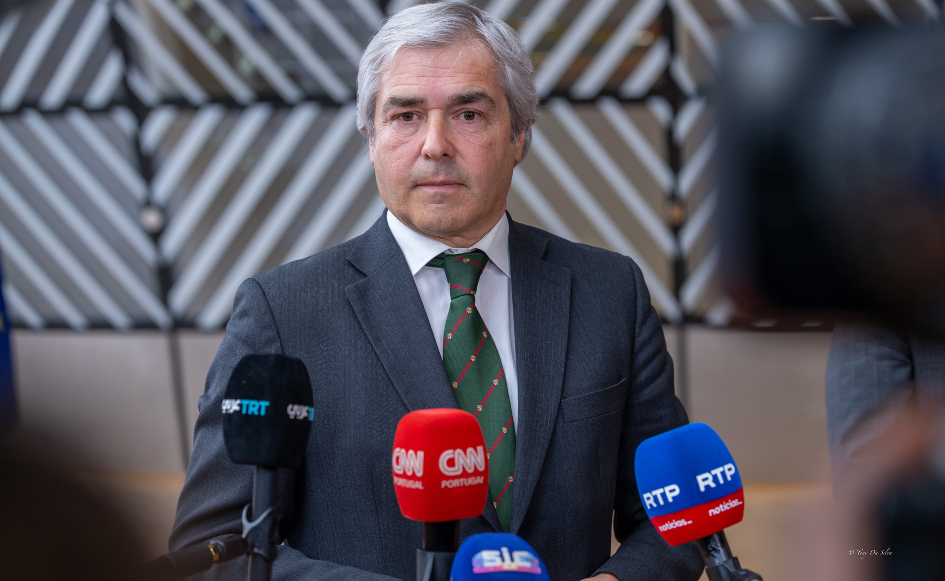 Ministro da Defesa português em conferência de imprensa, rodeado por microfones de vários órgãos de comunicação social, durante uma reunião da União Europeia em Bruxelas.