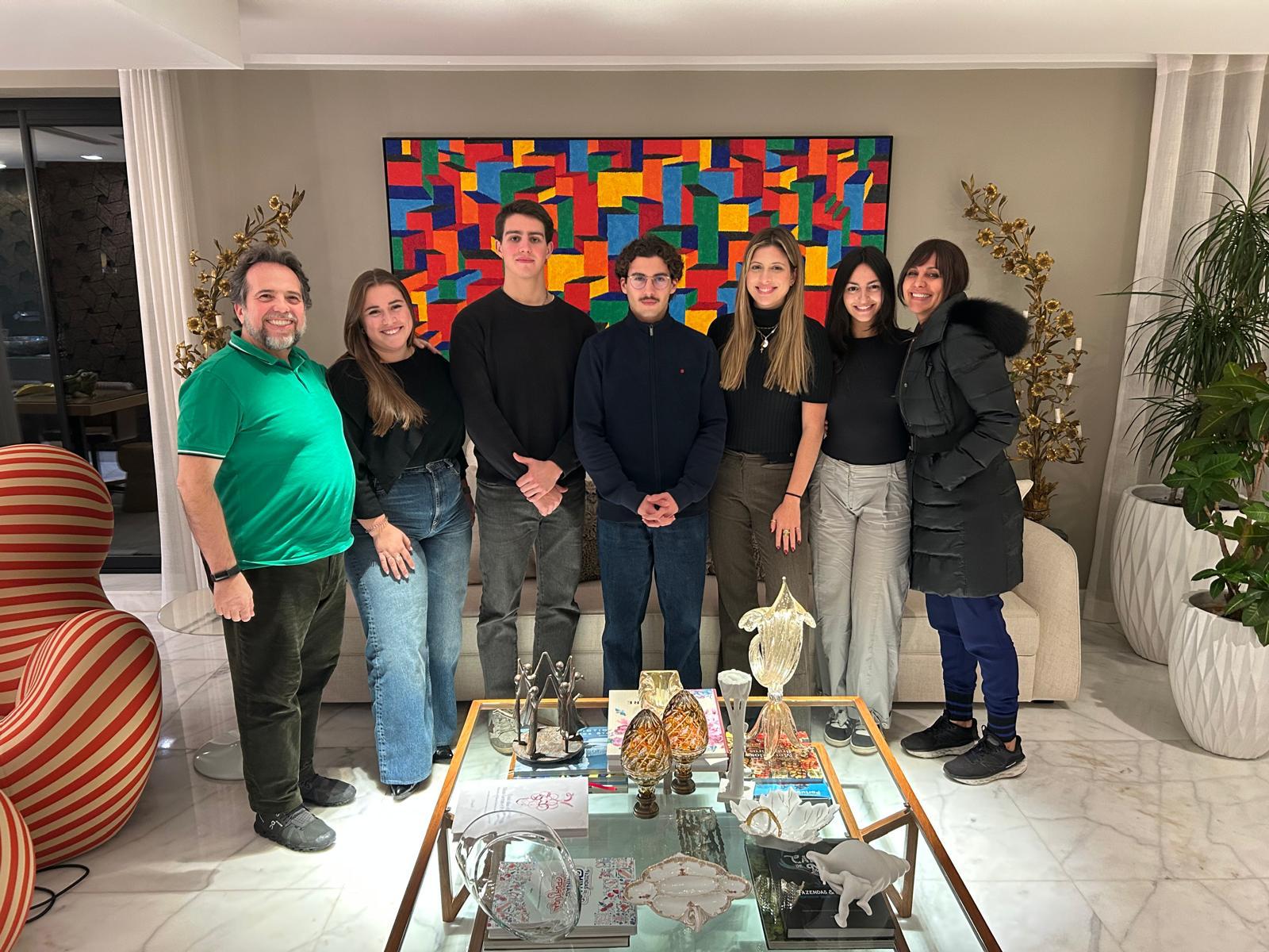 Sete jovens adultos em pé, lado a lado, num ambiente interior moderno e iluminado, com uma mesa de centro e uma pintura colorida ao fundo, simbolizando o início das atividades do Comité Câmara Jovem da Câmara de Comércio e Indústria Luso-Brasileira.