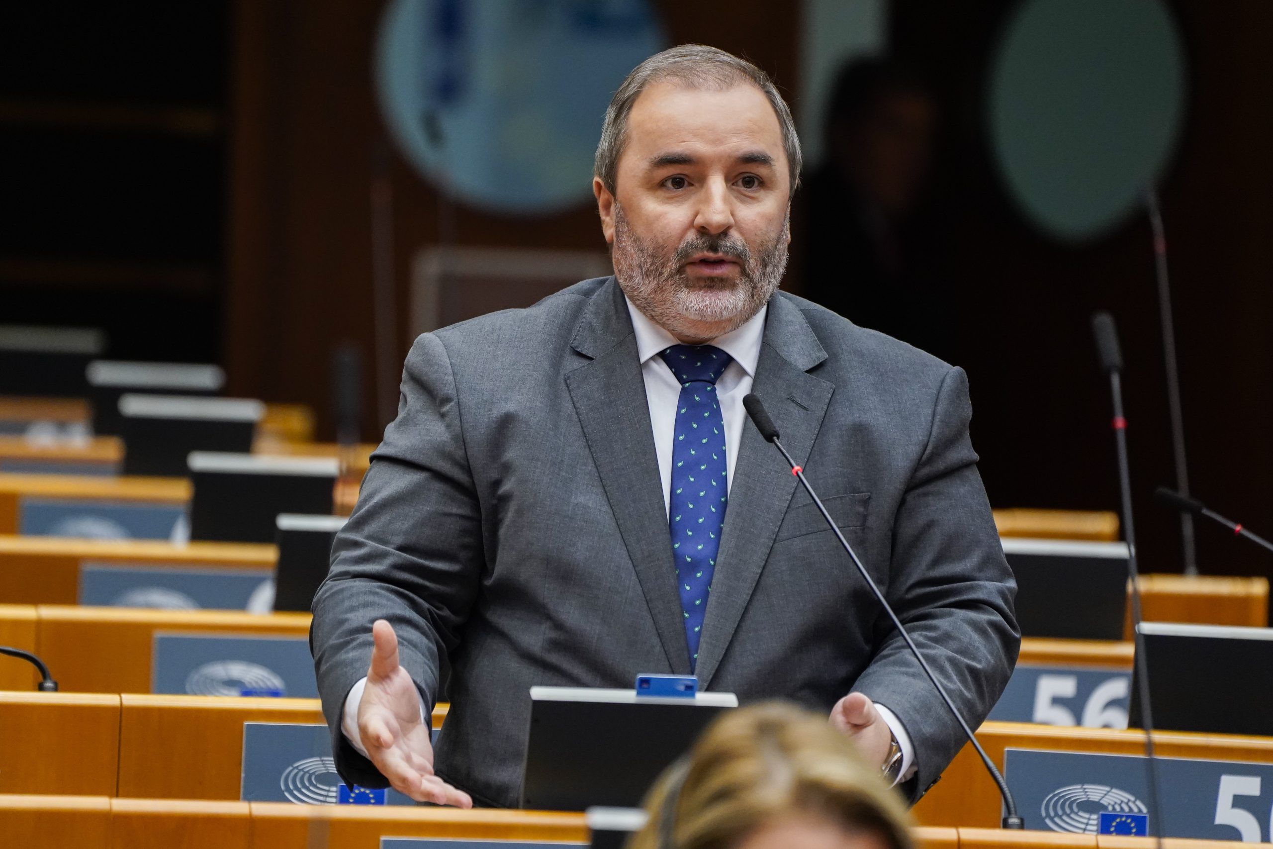 Eurodeputado a discursar numa sala do Parlamento Europeu, de pé junto ao microfone, com lugares vazios ao fundo, ilustrando uma intervenção formal em contexto parlamentar.