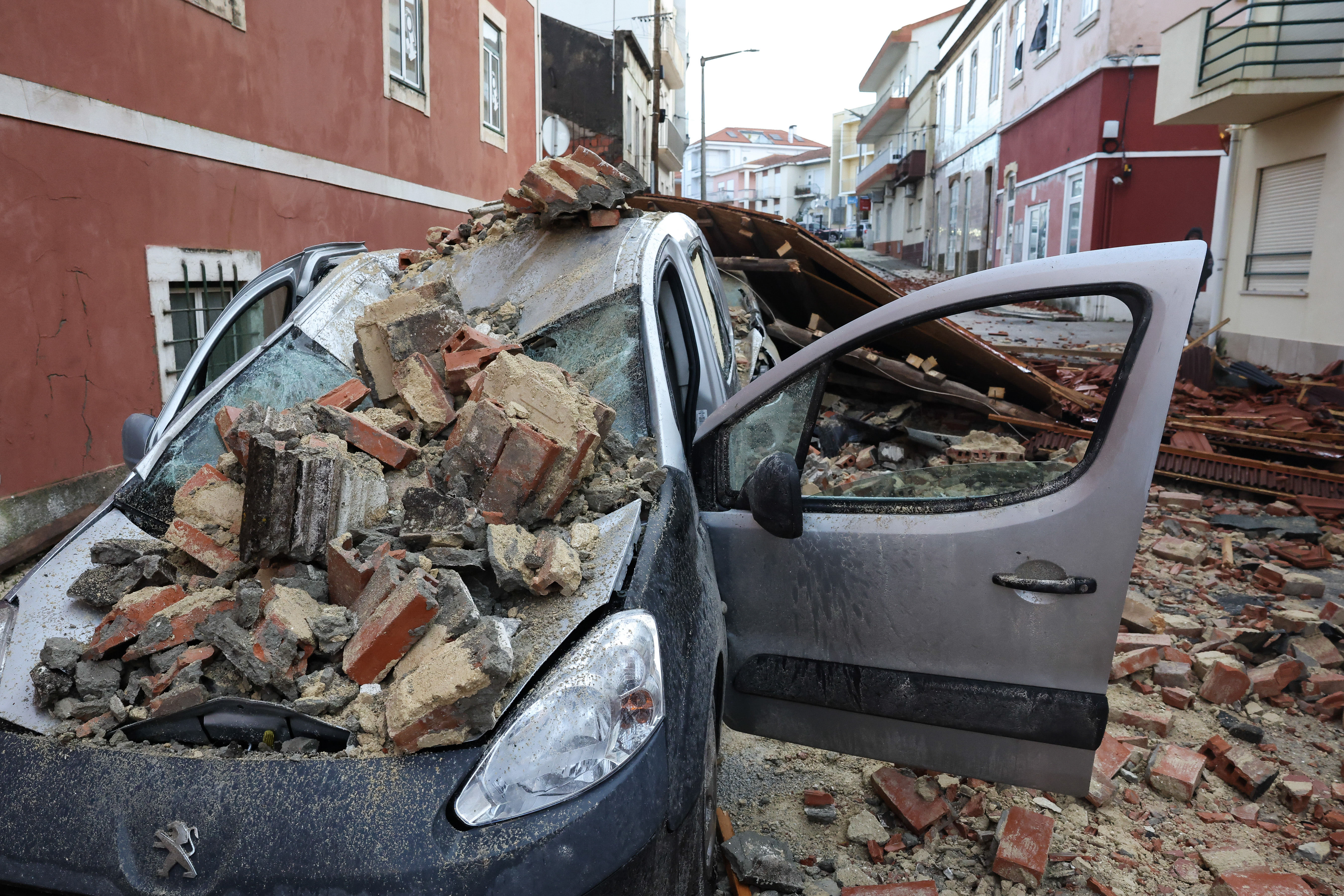 Carro esmagado por tijolos e escombros numa rua urbana, ilustrando os danos causados pelo mau tempo em Portugal.