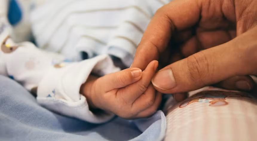 Mão de adulto a segurar suavemente a mão de um bebé, simbolizando ligação e proteção familiar.