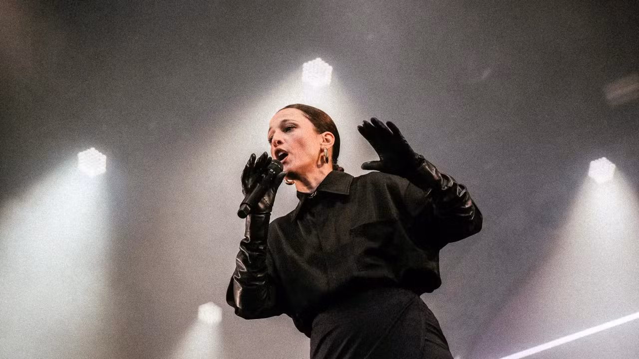 Um artista no palco, vestindo roupas pretas e luvas, cantando em um microfone com luzes brilhantes ao fundo.