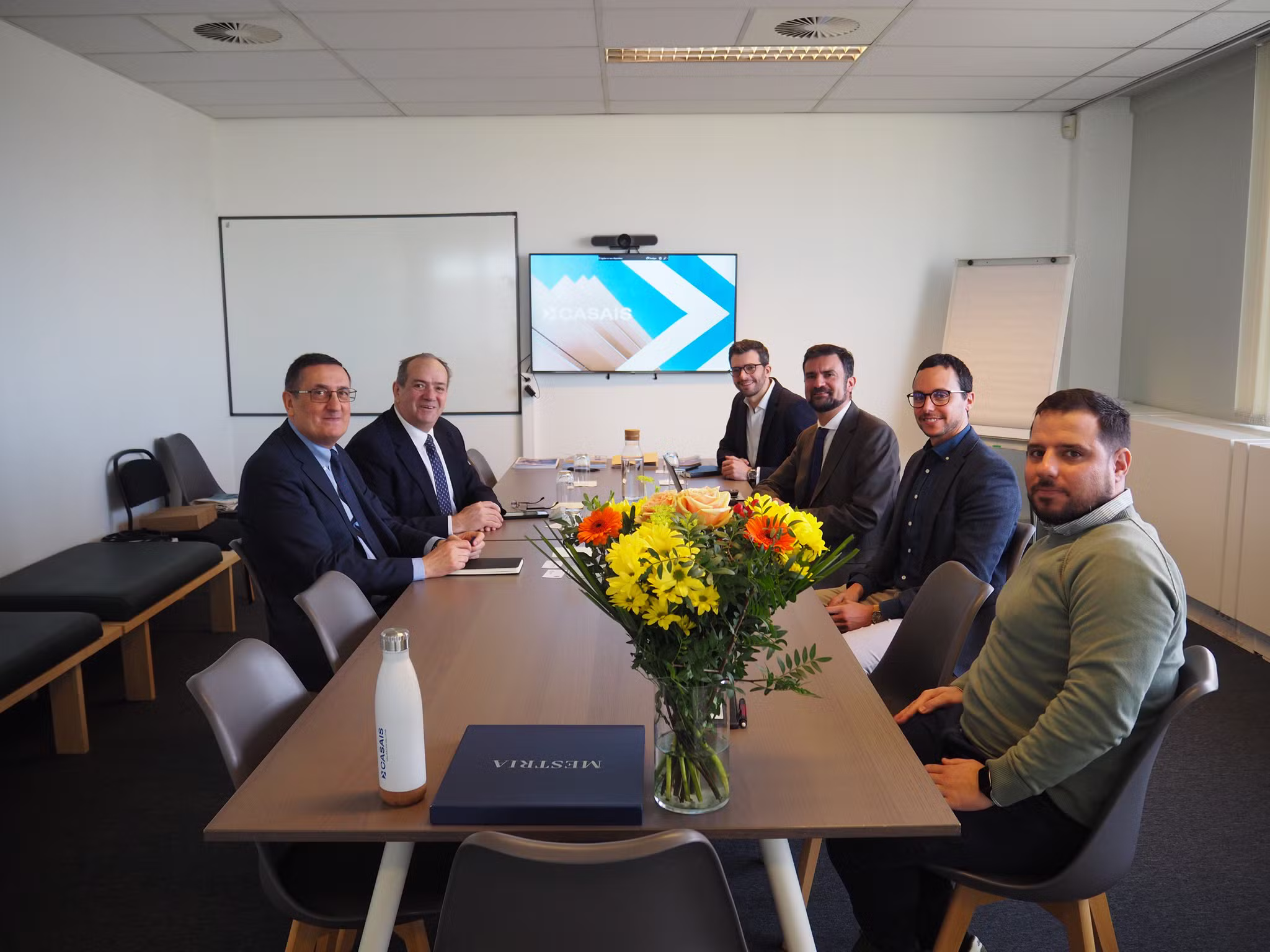 Seis pessoas sentadas à volta de uma mesa de reuniões, com flores no centro e um ecrã de apresentação ao fundo, durante uma reunião formal nos escritórios da Casais Bélgica.