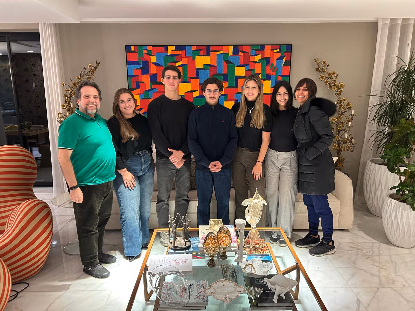 Sete jovens adultos em pé, lado a lado, num ambiente interior moderno e iluminado, com uma mesa de centro e uma pintura colorida ao fundo, simbolizando o início das atividades do Comité Câmara Jovem da Câmara de Comércio e Indústria Luso-Brasileira.
